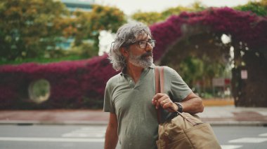 Gri saçlı ve sakallı, samimi, orta yaşlı bir adam yaya geçidinden karşıya geçiyor. Gözlüklü olgun bir beyefendi yaya geçidinden karşıya geçerken arkasına bakıyor.