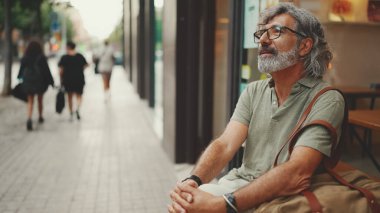 Thoughtful middle-aged man with gray hair and beard, wearing casual clothes, sits in street cafe. Mature gentleman in eyeglasses
