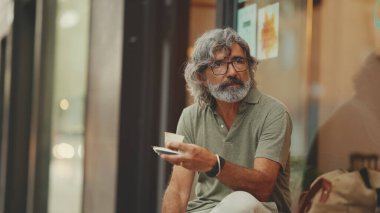 Gri saçlı, sakallı, sıradan kıyafetler giyen düşünceli, orta yaşlı bir adam sokak kafesinde oturuyor. Gözlüklü olgun beyefendi aromalı kahve içer.