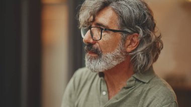 Thoughtful middle-aged man with gray hair and beard, wearing casual clothes, sits in street cafe. Mature gentleman in eyeglasses