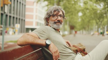 Gri saçlı, sakallı, sıradan kıyafetler giyen orta yaşlı bir adam bankta oturuyor. Gözlüklü olgun beyefendi bankta dinleniyor.