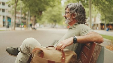 Gri saçlı, sakallı, sıradan kıyafetler giyen orta yaşlı bir adam bankta oturuyor. Gözlüklü olgun beyefendi bankta dinleniyor.