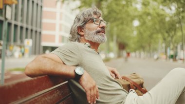 Gri saçlı, sakallı, sıradan kıyafetler giyen orta yaşlı bir adam bankta oturuyor. Gözlüklü olgun beyefendi bankta dinleniyor.
