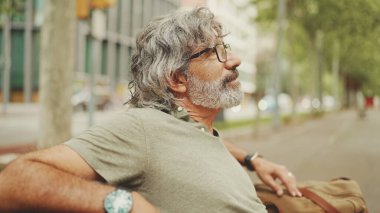 Gri saçlı, sakallı, sıradan kıyafetler giyen orta yaşlı bir adam bankta oturuyor. Gözlüklü olgun beyefendi bankta dinleniyor.