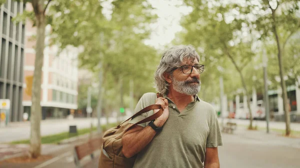 Gri saçlı, sakallı orta yaşlı bir adam yürüyor ve etrafına bakıyor. Omzunda çanta olan gözlüklü olgun bir beyefendi şehir manzarası arka planında meydanda yürüyor.