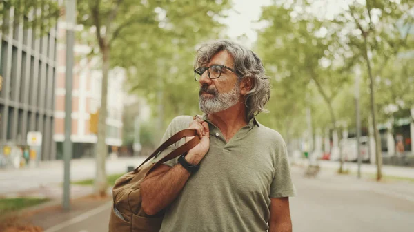 Gri saçlı, sakallı orta yaşlı bir adam yürüyor ve etrafına bakıyor. Omzunda çanta olan gözlüklü olgun bir beyefendi şehir manzarası arka planında meydanda yürüyor.