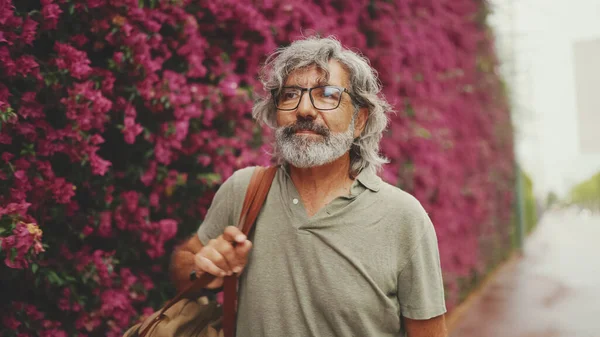 Dost canlısı, orta yaşlı, gri saçlı ve sakallı bir adam, arka planda çiçek açan mor çiçeklerle duvarın yanında yürüyor.