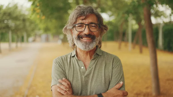Gri saçlı, sakallı, sıradan kıyafetler giyen, dost canlısı, orta yaşlı bir adam kameraya bakıyor. Gözlüklü olgun beyefendi kameraya gülümsüyor.