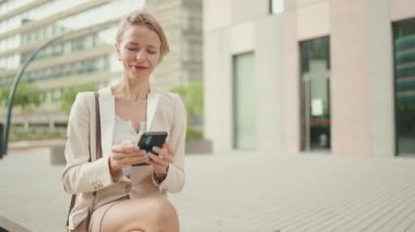 Smiling woman with blond hair wearing beige suit using cellphone while sitting outside on the street