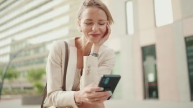 Smiling woman with blond hair wearing beige suit using cellphone while sitting outside on the street