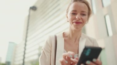Smiling woman with blond hair wearing beige suit using cellphone while sitting outside on the street