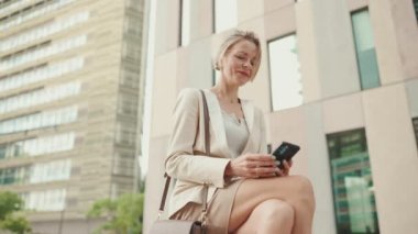 Smiling woman with blond hair wearing beige suit using mobile cellphone sitting outside on the street