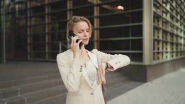 Businesswoman leaves the business center, talking on the cellphone, looking at her watch