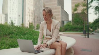 Smiling businesswoman with blond hair wearing beige suit is using pc laptop outside