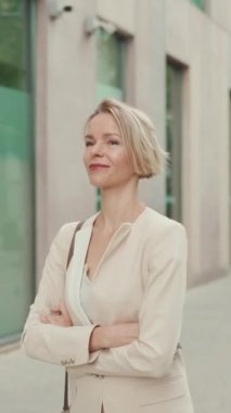 VERTICAL VIDEO: Smiling woman with blond hair wearing beige suit walks down the street and looks around
