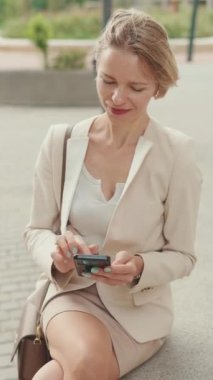 VERTICAL VIDEO: Smiling woman with blond hair wearing beige suit using mobile cellphone sitting outside on the street