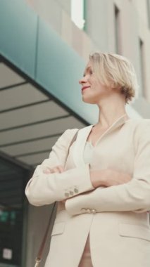 VERTICAL VIDEO: Smiling woman with blond hair wearing beige suit walks down the street and looks around
