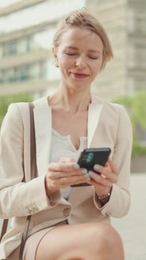 VERTICAL VIDEO: Smiling woman with blond hair wearing beige suit using mobile cellphone sitting outside on the street