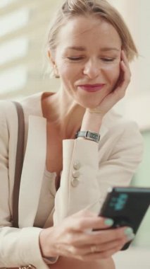 VERTICAL VIDEO: Smiling woman with blond hair wearing beige suit using mobile cellphone sitting outside on the street