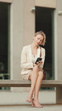 VERTICAL VIDEO: Businesswoman with blond hair using cellphone outside on the street