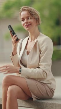 VERTICAL VIDEO: Businesswoman with blond hair talking on cellphone, looking at wrist watch
