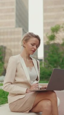 VERTICAL VIDEO: Businesswoman with blond hair wearing beige suit is using pc laptop outside