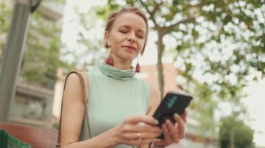 Beautiful smiling woman with short blond hair in casual clothes sits on bench, uses cellphone