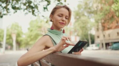 Beautiful smiling woman with short blond hair in casual clothes sits on bench, uses cellphone