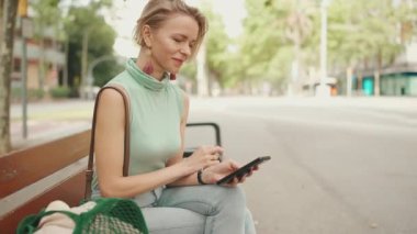 Beautiful smiling woman with short blond hair in casual clothes sits on bench, uses cellphone