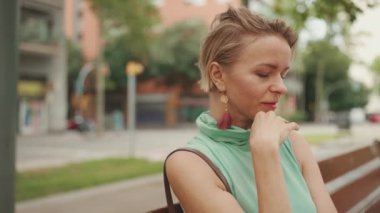 Beautiful woman with short blond hair in casual clothes sits on bench, smiles, enjoys warm summer day