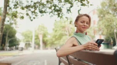 Beautiful smiling woman with short blond hair in casual clothes sits on bench, uses cellphone