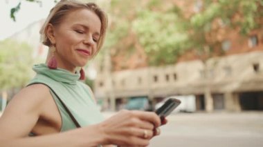 Beautiful smiling woman with short blond hair in casual clothes sits on bench, uses cellphone