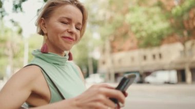 Beautiful smiling woman with short blond hair in casual clothes sits on bench, uses cellphone