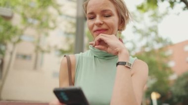 Beautiful happy woman with short blond hair in casual clothes sits on bench, uses cellphone