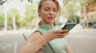 Beautiful smiling woman with short blond hair in casual clothes sits on bench, sends voice message on cellphone