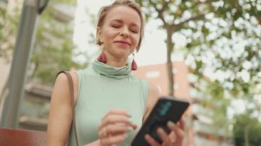 Beautiful smiling woman with short blond hair in casual clothes sits on bench, uses cellphone