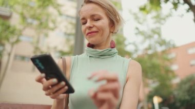Beautiful smiling woman with short blond hair in casual clothes sits on bench, uses cellphone