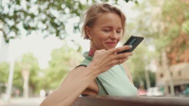 Beautiful happy smiling woman with short blond hair in casual clothes sits on bench, sends voice message on cellphone