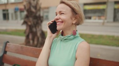 Close up, beautiful happy smiling woman with short blond hair in casual wear sits on bench, talking on cellphone