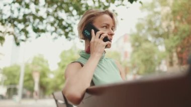 Beautiful happy smiling woman with short blond hair in casual wear sits on bench, talking on cellphone