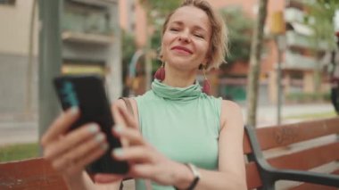 Beautiful happy smiling woman with short blond hair in casual wear sits on bench, makes video call on cellphone