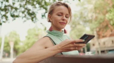 Beautiful smiling woman with short blond hair in casual clothes sits on bench, sends voice message on cellphone
