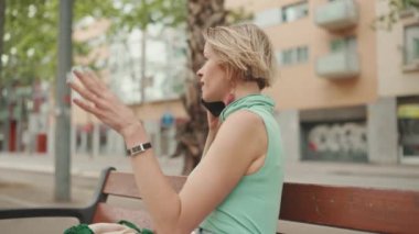 Profile beautiful happy smiling woman with short blond hair in casual wear sits on bench, talking on cellphone