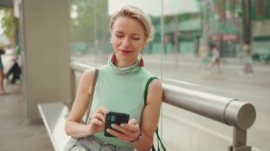 Smiling beautiful woman with short blond hair in casual clothes sits at public transport stop, uses mobile phone