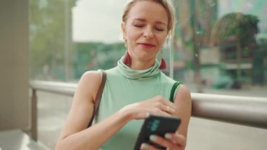 Smiling beautiful woman with short blond hair in casual clothes sits at public transport stop, uses mobile phone