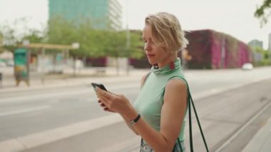 Beautiful woman with short blond hair in casual clothes stands at tram stop, uses phone