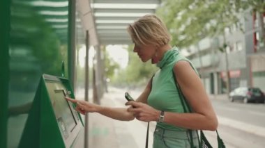 Beautiful woman with short blond hair in casual clothes stands at tram stop, buys ticket to travel on public transport
