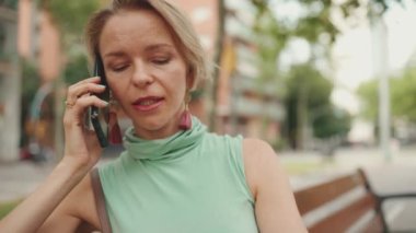 Close up, beautiful happy smiling woman with short blond hair in casual wear sits on bench, talking on cellphone