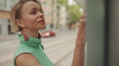 Close up, beautiful woman with short blond hair in casual clothes stands at tram stop, looks at the public transport timetable on the information board