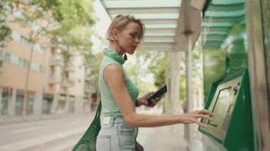 Beautiful woman with short blond hair in casual clothes stands at tram stop, buys ticket to travel on public transport